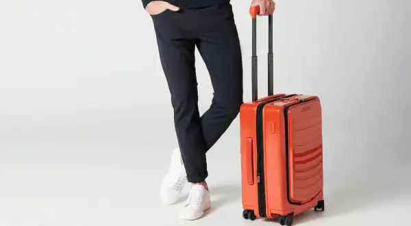 Mężczyzna w eleganckim stroju stoi obok pomarażowej walizki. Różne rozmiary walizek dostępne są dla każdego podróżnika.