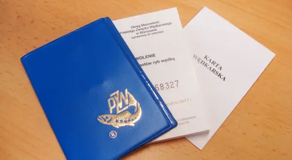 Niebieska książeczka z logo PZW i dokumenty "Karta Wędkarska" na drewnianym tle.