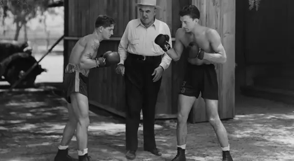Two boxers spar while a trainer watches. The boxer on the left has a tattoo. This scene evokes the spirit of legendary boxer James J. Jeffries.