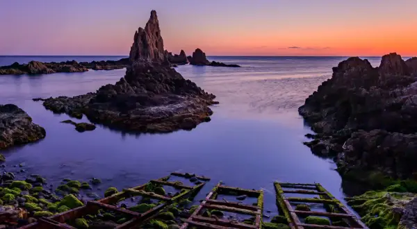 Rocas afiladas emergen del mar sereno bajo un cielo crepuscular. La **sierra Cabo de Gata** se revela en su esplendor, con restos oxidados de estructuras en primer plano.