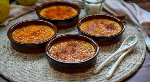 Vier Crema Catalana Schalen mit karamellisierter Zuckerkruste, bereit für das Crema Catalana Rezept. Zitronen und Löffel liegen daneben.