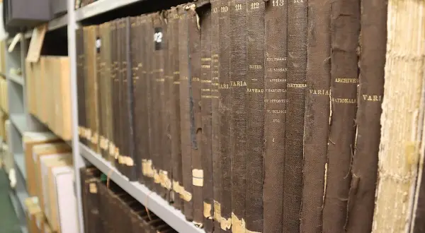 Rangées de vieux registres reliés en tissu brun dans les archives départementales 85. Les titres sont partiellement lisibles.