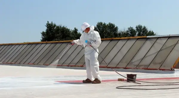Pracownik w białym kombinezonie natryskuje nowoczesne materiały na dach płaski EPDM, porównując je z papą termozgrzewalną.