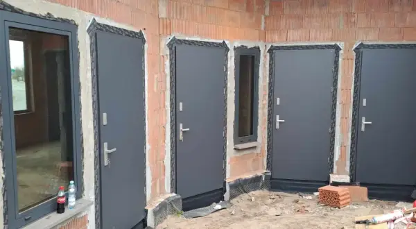Nowe, szare drzwi zewnętrzne i okno w budowie. Widać ceglane ściany i materiały budowlane.