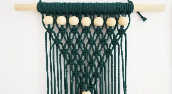 Tenture murale en macramé vert foncé avec perles en bois sur une tige en bois.