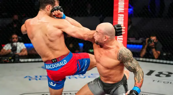 En MMA-fighter med tatueringar på armen, likt Gleison Tibau, tar emot en spark i ansiktet.