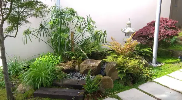 Un sereno mini jardín japonés en casa, con cascada de agua, rocas, plantas exuberantes y una linterna de piedra.