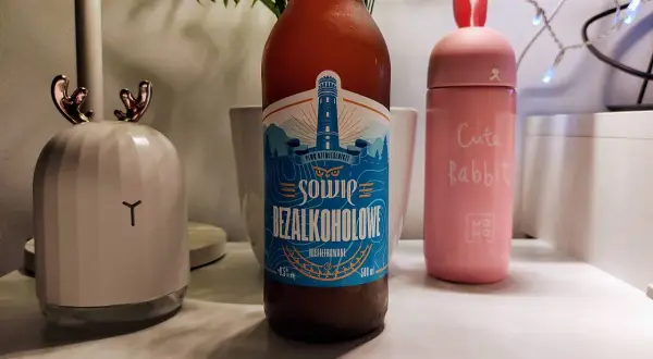 Butelka piwa Sowie Bezalkoholowe 0,5% obok różowego termosu z uszami królika i lampki w kształcie jelonka.