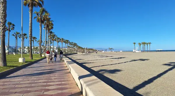 Paseo marítimo con palmeras en el **parque Roquetas de Mar**. Gente camina por la senda adoquinada junto a la playa de arena bajo un cielo azul.