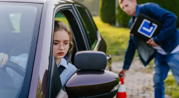 Dziewczyna w okularach za kierownicą samochodu, instruktor z tablicą "L" obok pachołka.