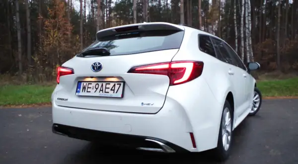 Biała Toyota Corolla 1.8 Hybrid na tle jesiennego lasu. Widok z tyłu, z podświetlonymi światłami.