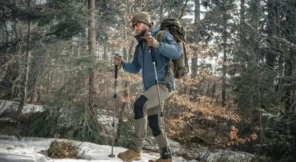 Mężczyzna w czapce i okularach przeciwsłonecznych idzie przez zaśnieżony las, wspierając się na kijach trekkingowych.