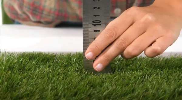 Mano mide la altura del césped artificial con una regla metálica. El césped es verde y denso.