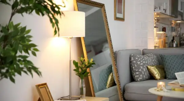 Ein großer Spiegel und eine Stehlampe machen die dunkle Wohnung heller. Ein Sofa mit Kissen und ein kleiner Tisch mit Kerze sind zu sehen.