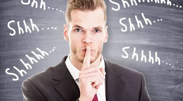 Uomo d'affari con dito sulle labbra, circondato da "Shhh..." scritti sulla lavagna. Il silenzio assenso significato è chiaro: non parlare.