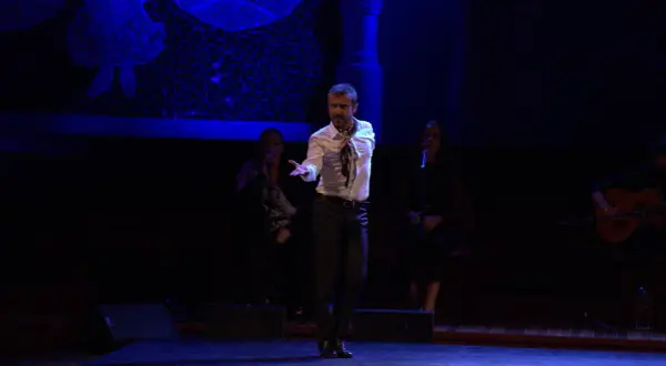 Un bailaor flamenco español actual, con camisa blanca y pañuelo, baila en el escenario. A su lado, dos cantantes y un guitarrista.