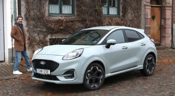 Man står bredvid Fords minsta modell, en ljusgrå Puma, framför ett gammalt hus med murgröna.