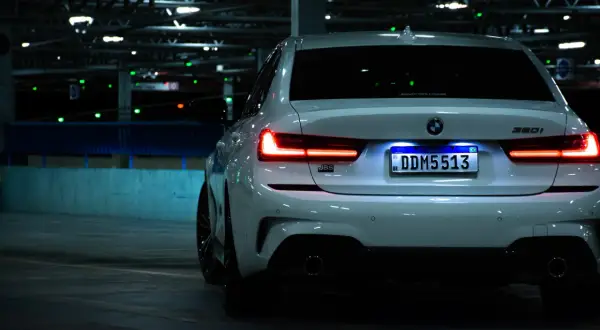 Vue arrière d'une BMW blanche dans un parking sombre. L'éclairage de la plaque d'immatriculation est bien visible, soulignant son caractère obligatoire.