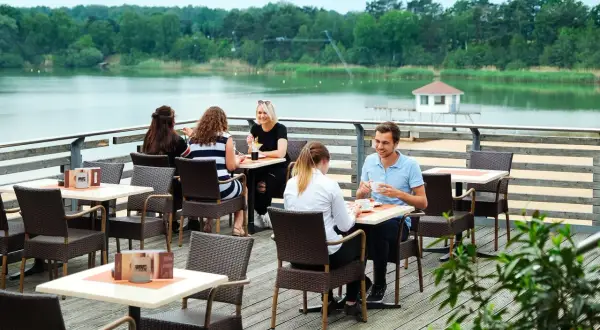 Gemütliches Restaurant am Wasser mit Gästen, die Kaffee und Getränke genießen. Im Hintergrund ein ruhiger See und grüne Bäume.