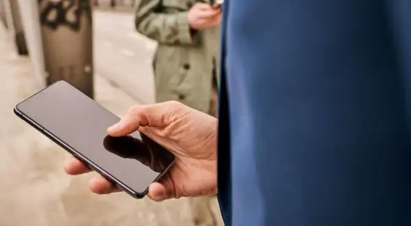 Mężczyzna trzyma smartfon, zastanawiając się, jak skonfigurować telefon. W tle druga osoba również korzysta z telefonu.