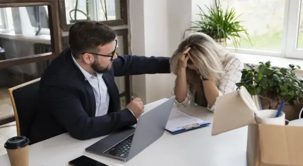 Mężczyzna pociesza kobietę przy biurku, na którym leży laptop i dokumenty.