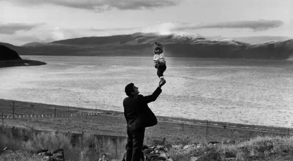 Photographie artistique : un homme soulève un bébé souriant au-dessus d'un lac, avec des montagnes en arrière-plan.