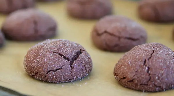 Mörka, sprickiga nutella kakor täckta med socker ligger på ett bakplåtspapper.
