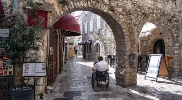 Wąska, brukowana uliczka w **Budva stare miasto**. Mężczyzna na wózku inwalidzkim przejeżdża pod kamiennym łukiem. Po bokach sklepy i restauracje.