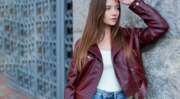 Woman in a burgundy leather jacket and blue jeans leans against a stone wall.