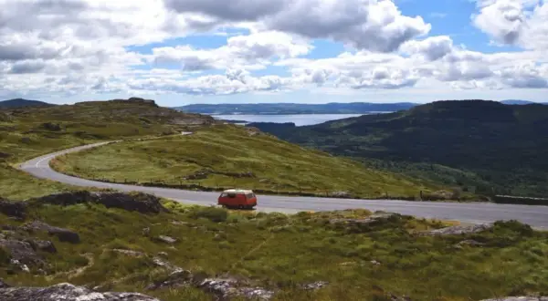 Eine malerische England Landschaft mit einer kurvigen Straße, einem roten Campervan und einem See in der Ferne unter bewölktem Himmel.