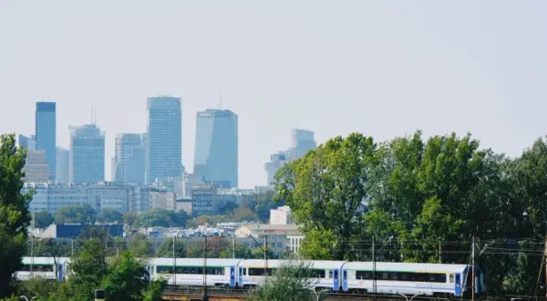 Pociąg PKP jedzie przez zielone drzewa z widokiem na warszawskie wieżowce. Szukasz tanich biletów PKP Warszawa - Kraków?