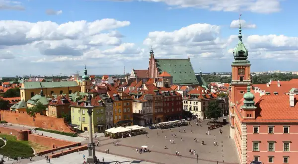 Widok na Zamek Królewski i Kolumnę Zygmunta III Wazy na placu w warszawskiej starówce. Tłumy spacerują po bruku, a słońce przebija się przez chmury.