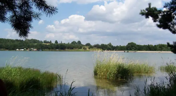 Spokojny zalew Dźibice otoczony zielenią, z kempingiem na brzegu i chmurami na niebie.