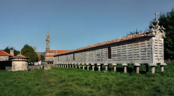 El horreo de Carnota, una estructura de piedra sobre pilares, se alza majestuoso ante una iglesia y un cielo azul.