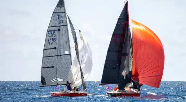 Due barche a vela competono in mare aperto. La barca a sinistra ha una vela grigia con numeri e simboli, mentre quella a destra sfoggia vele nere e arancioni. Lo strallo, elemento cruciale per la stabilità, è ben visibile.