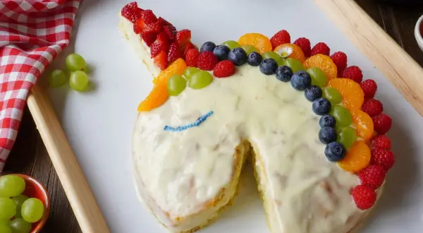 Ein fantasievoller Kuchen ohne Zucker in Einhornform, verziert mit bunten Früchten wie Erdbeeren, Blaubeeren und Mandarinen.
