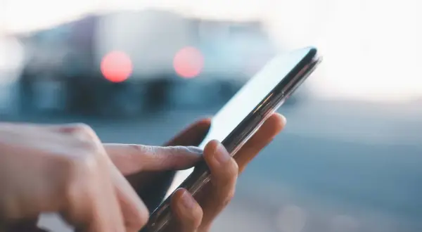 Dłoń dotyka ekranu smartfona, szukając informacji, jak przywrócić ustawienia fabryczne w telefonie. W tle rozmyte światła.