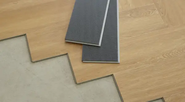 Panele winylowe w trakcie układania na betonowej podłodze. Dwa szare panele leżą na drewnopodobnych deskach.