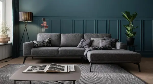 Ein moderner Wohnbereich mit einem grauen Ecksofa, das auf einem gemusterten Teppich steht. Ein Couchtisch mit einem offenen Buch und eine Stehlampe vervollständigen das Bild.