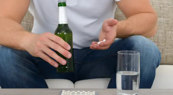 Mężczyzna trzyma butelkę piwa i garść tabletek, obok stoi szklanka wody. Czy to ibuprofen i alkohol?