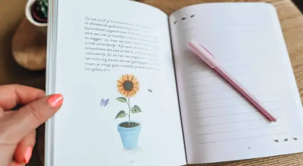 Een open dagboek met een zonnebloem en vlinder illustratie. Een roze pen ligt op de rechterpagina, klaar om gedachten uit je hoofd op te schrijven.