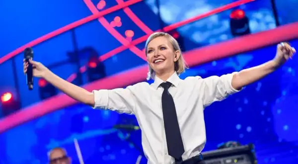 Kasia Stankiewicz na scenie, w białej koszuli i krawacie, śpiewa z mikrofonem. W tle niebieska scenografia z czerwonymi elementami, przypominająca orła cień.