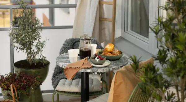 En balkong med tropisk inredning, kaffe, bullar och bär. Gröna växter och kuddar skapar en ombonad känsla.