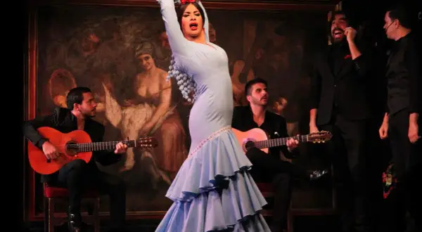 Bailarina de flamenco con vestido azul, guitarristas y cantantes en un escenario. Una escena que evoca el origen de la palabra flamenco.