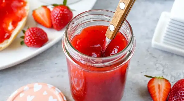 Ein Glas mit leuchtend roter Erdbeermarmelade, die mit einem Holzlöffel umgerührt wird. Daneben frische Erdbeeren und ein halbes Brötchen mit Marmelade – Erdbeermarmelade mit Pfiff für den perfekten Start in den Tag.