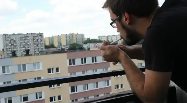 Mężczyzna zapala papierosa na balkonie, patrząc na blokowisko. Czy można palić na balkonie? To pytanie zadaje sobie wielu mieszkańców.