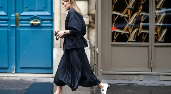 Elegancka kobieta w czarnej, plisowanej spódnicy i marynarce, spacerująca po ulicy. Stylizacje z plisowaną spódnicą są zawsze modne.