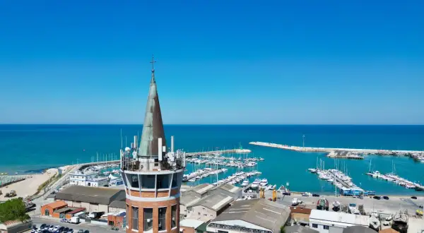 Torre con antenne domina il porto civitanova, pieno di barche e con il mare azzurro sullo sfondo.