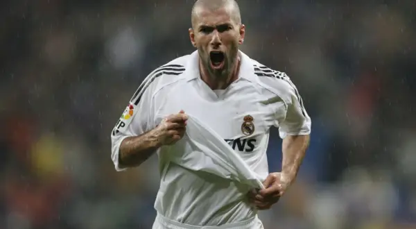 Zinedine Zidane w białej koszulce Realu Madryt, z podniesioną koszulką, krzyczy radośnie do kibiców.