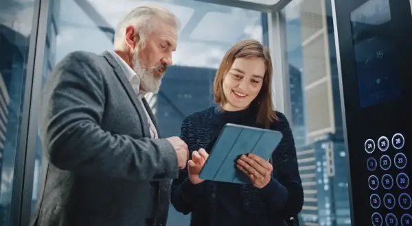 Starszy mężczyzna i młoda kobieta omawiają projekt na tablecie w windzie. To idealny moment na ich elevator pitch.